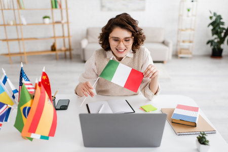 joyful language teacher showing Italian flag during online lesson on laptop near notebooks and smartphoneの写真素材