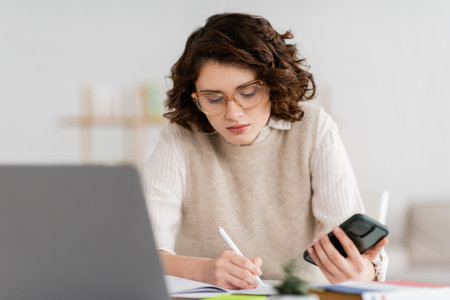 pretty student in glasses holding smartphone while taking notes near blurred laptop on deskの写真素材