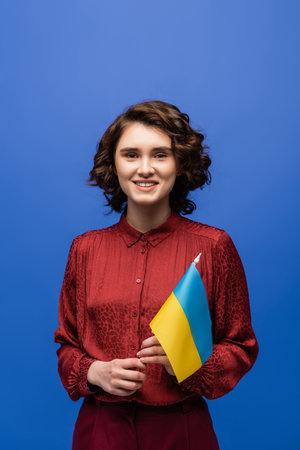 happy language teacher holding flag of Ukraine and smiling isolated on blueの写真素材