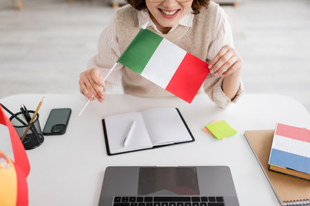 cropped view of smiling language teacher holding Italian flag near devices and notebooks on table at homeの写真素材