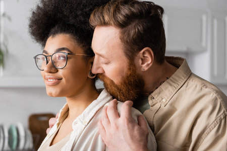 Bearded man hugging smiling african american girlfriend at homeの写真素材