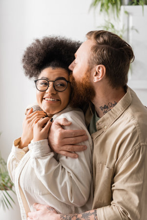 Bearded man kissing and hugging smiling african american girlfriend at homeの写真素材