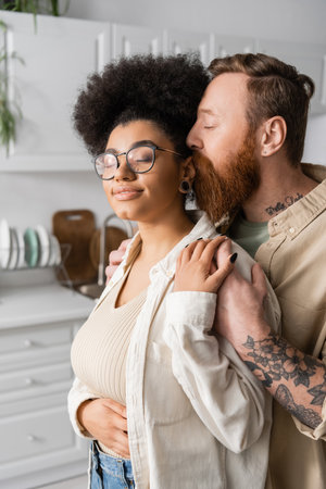 Tattooed man hugging and kissing smiling african american girlfriend at homeの写真素材