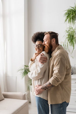 Tattooed man hugging cheerful african american girlfriend in eyeglasses at homeの写真素材