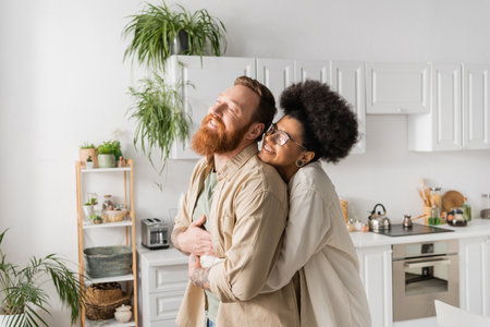 Smiling african american woman in eyeglasses hugging boyfriend at homeの写真素材