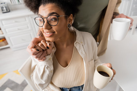 Blurred man holding hand of smiling african american girlfriend with coffee cup in kitchenの写真素材