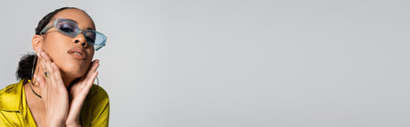 pretty african american woman in blue sunglasses and stylish shirt touching face isolated on grey, bannerの写真素材