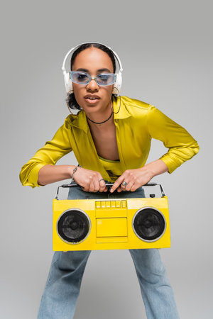 stylish african american woman listening music in wireless headphones while holding boombox isolated on greyの写真素材