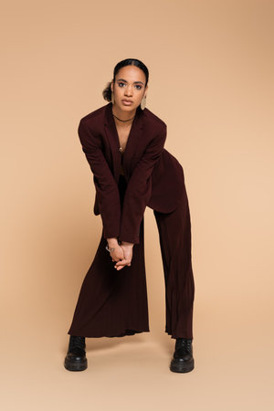 full length of young african american woman in maroon blazer and wide pants posing on beigeの写真素材