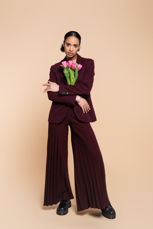 full length of well dressed african american woman in burgundy suit posing with pink tulips on beigeの写真素材