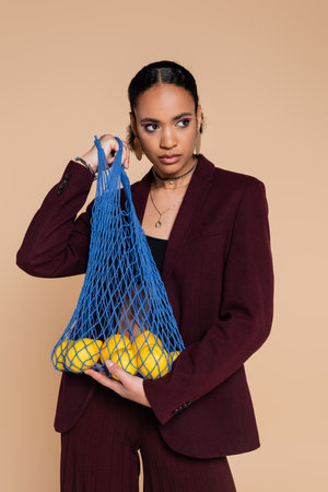 stylish african american woman in burgundy suit holding mesh bag with lemons isolated on beigeの写真素材