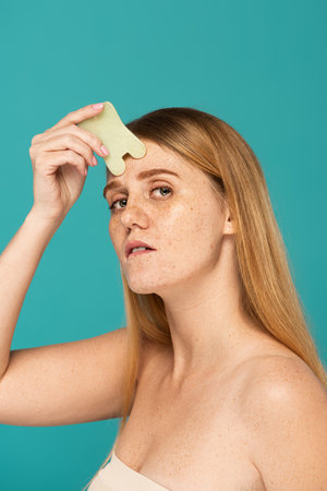 young and redhead woman with freckles using face scrapper isolated on turquoiseの写真素材