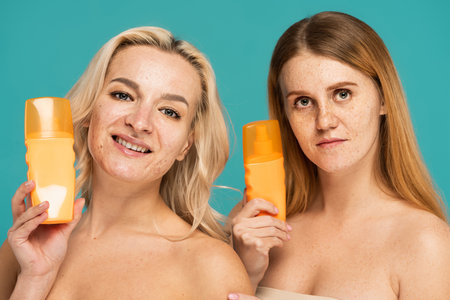 happy women with bare shoulders holding bottles with sunscreen isolated on turquoiseの写真素材