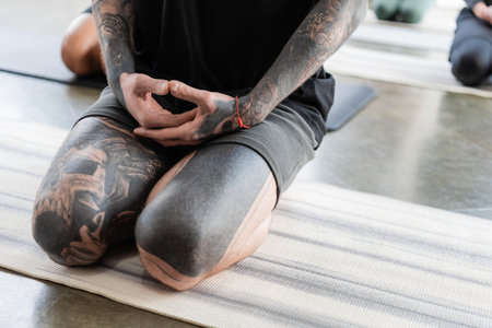 Cropped view of tattooed man sitting in Thunderbolt asana on yoga mat in studioの写真素材