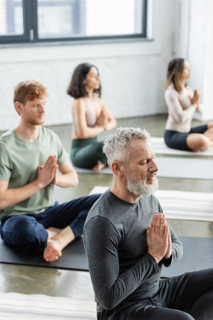 Mature man sitting in anjali mudra near blurred multiethnic group in yoga classの写真素材