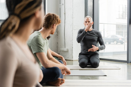 Middle aged coach explaining nostril breathing near blurred interracial group in yoga classの写真素材