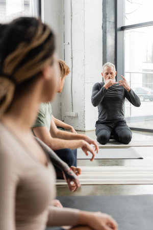Mature coach practicing nostril breathing near blurred interracial group in yoga classの写真素材
