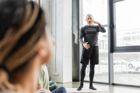 Mature coach talking and explaining asana to blurred interracial group in yoga classの写真素材