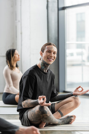Positive tattooed man looking at camera while doing gyan mudra in yoga studioの写真素材