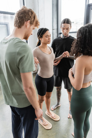 tattooed african american woman with closed eyes standing on sadhu nail board near people in yoga...