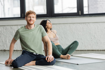 Smiling redhead man looking at camera near middle eastern woman in yoga studioの写真素材