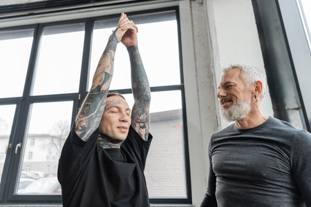 Smiling middle aged coach looking at tattooed man doing Crescent Lunge asana in yoga classの写真素材
