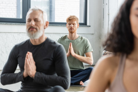 Redhead man meditating with anjali mudra near blurred people in yoga classの写真素材