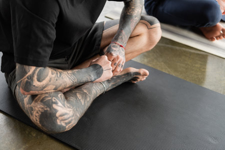Cropped view of tattooed man sitting in fire log asana on mat in yoga classの写真素材