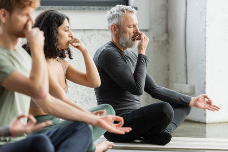 Mature man practicing nostril breathing and gyan mudra near interracial people in yoga studioの写真素材