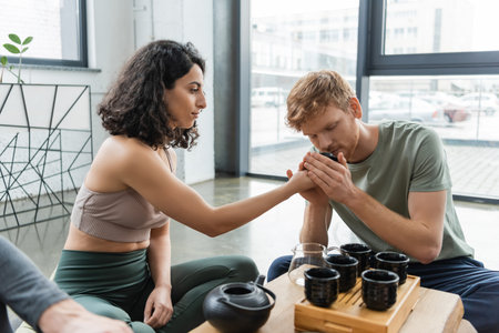 redhead man smelling puer tea near curly middle eastern woman near Chinese teapot and cupsの写真素材