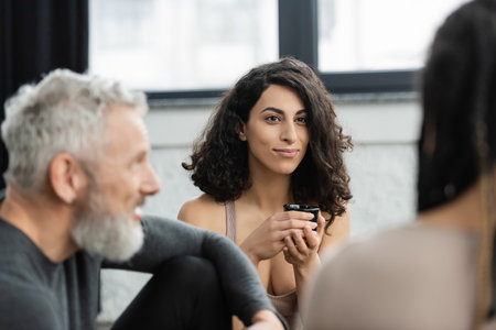 happy middle eastern woman with curly hair holding japanese cup during tea ceremony in yoga studioの写真素材