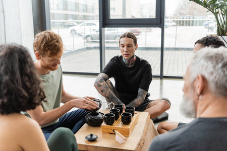 young man with tattoos sitting near interracial friends during tea ceremony in yoga studioの写真素材