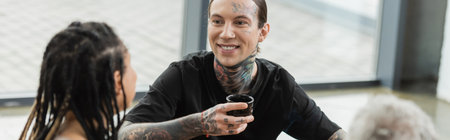 happy and tattooed man holding Chinese tea cup and talking with african american woman in yoga studio, bannerの写真素材