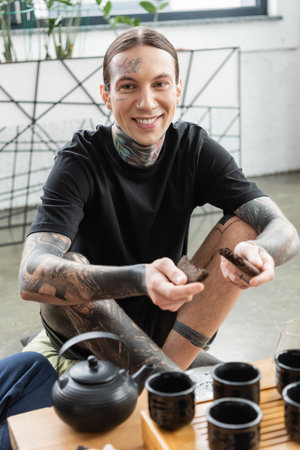 cheerful young man with tattoos breaking compressed puer tea near traditional Chinese tea pot and cupsの写真素材