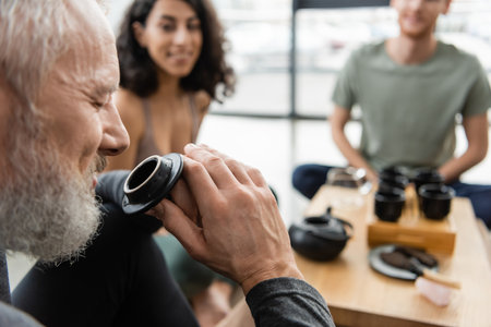 happy middle aged man with closed eyes enjoying aroma of puer tea near people on blurred backgroundの写真素材