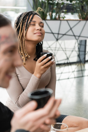 pleased african american woman with dreadlocks holding cup with puer tea near tattooed man on blurred foregroundの写真素材