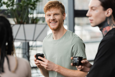 cheerful man with red hair holding japanese cup with puer tea near people in yoga studioの写真素材