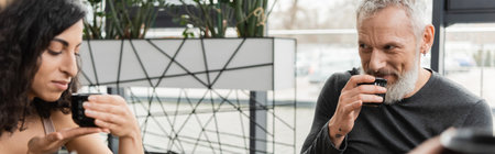 happy mature man drinking puer tea while looking at middle eastern woman in yoga studio, bannerの写真素材