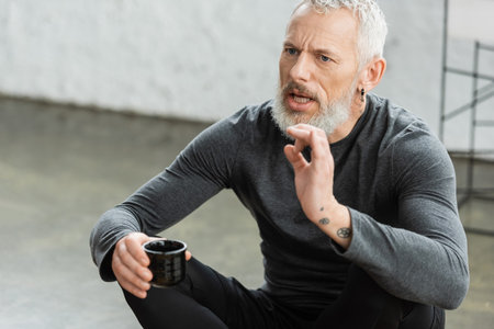 bearded middle aged man with tattoos holding Chinese cup with puer tea while talking in yoga studioの写真素材