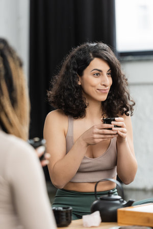 happy middle eastern woman holding Chinese cup during tea ceremony in yoga studioの写真素材