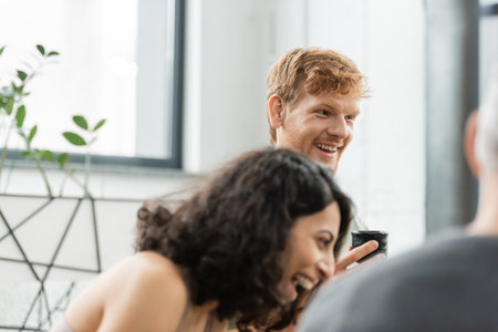 happy redhead man holding tea cup with puer and laughing near middle eastern woman in yoga studioの写真素材