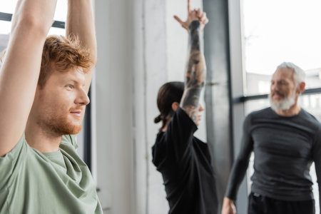 Smiling redhead man standing in Crescent Lunge asana near blurred group in yoga studioの写真素材