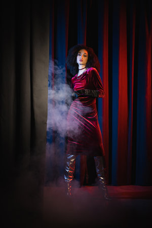 Young african american model in burgundy dress standing on red background with blue light and smokeの写真素材