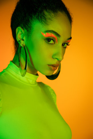 Fashionable african american model with neon eyeliner looking at camera on orange backgroundの写真素材