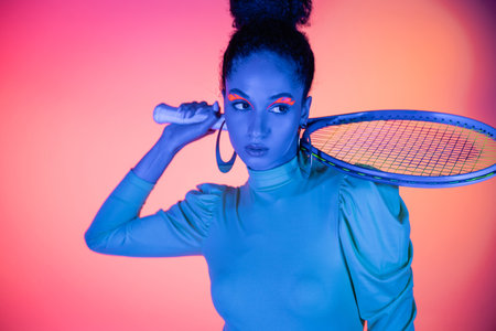 Fashionable african american woman with neon eyeliner holding tennis racket on pink backgroundの写真素材
