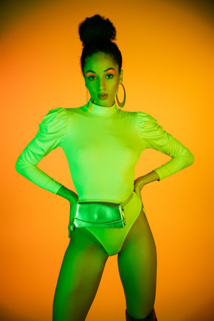 Trendy african american model with neon eyeliner and bodysuit holding hands on hips on orange backgroundの写真素材