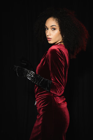 Trendy african american woman in velvet burgundy dress and glove looking at camera isolated on blackの写真素材