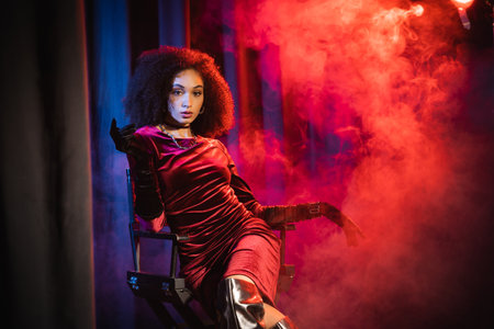 african american woman in velvet dress and gloves looking at camera while sitting on chair on background with red light and smokeの写真素材