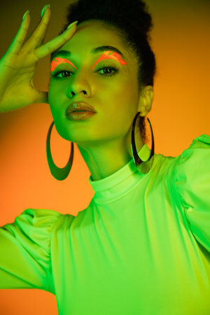 Portrait of fashionable african american woman with neon visage standing on orange backgroundの写真素材