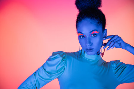 Portrait of stylish african american woman with neon visage touching earring on light pink backgroundの写真素材